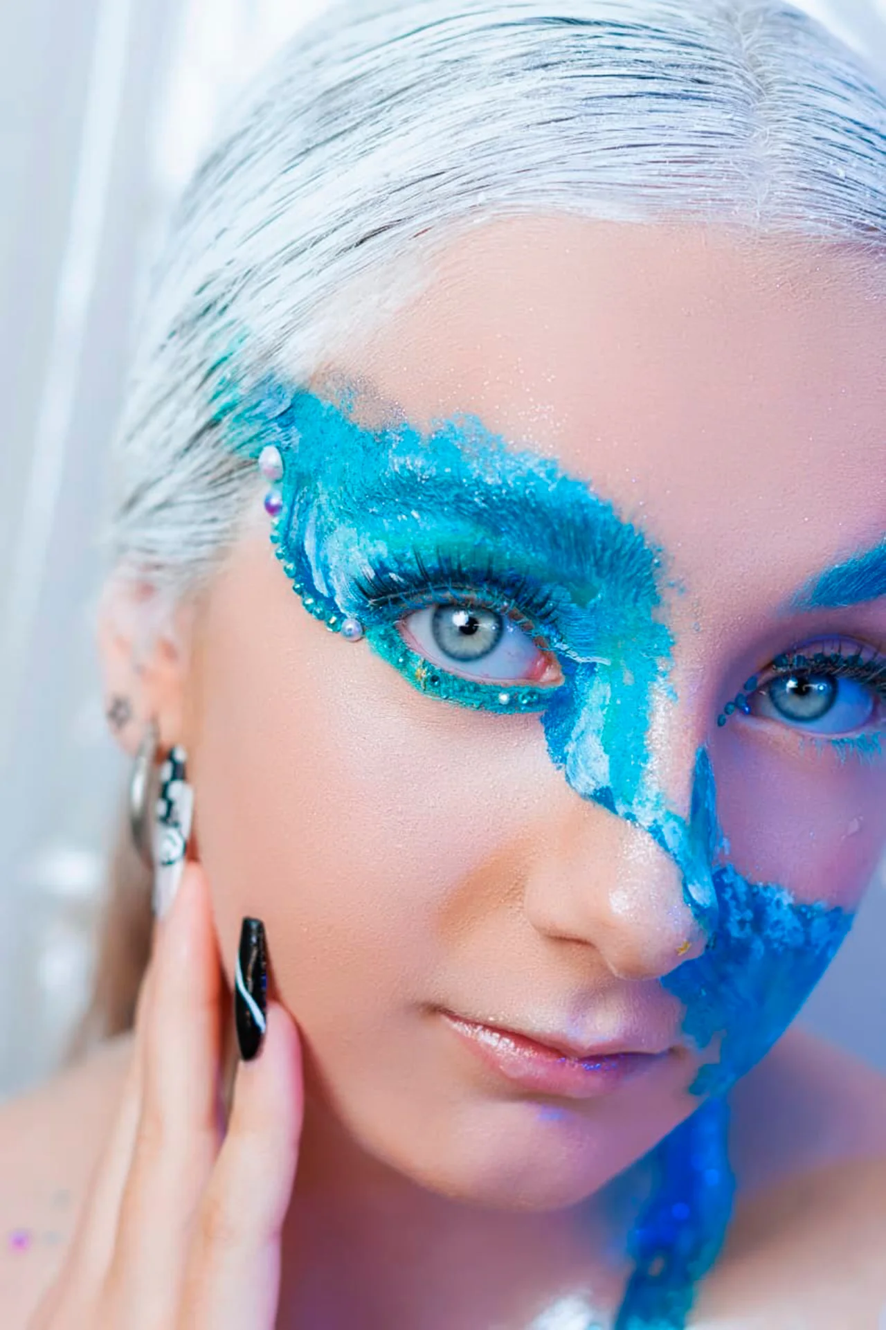 Primer plano de una persona con un maquillaje facial artístico de belleza. Sus ojos están cubiertos por un diseño vibrante de azul turquesa y blanco que parece una ola o sal texturizada, extendiéndose en forma de máscara alada. El borde del maquillaje está adornado con gemas y cristales azules y blancos. Su cabello es corto y de color blanco escarchado. Tiene ojos azules claros que miran directamente a la cámara. Su mano derecha, con uñas pintadas de negro con diseños blancos abstractos, toca su mejilla. Se ven varios pendientes de plata en su oreja. El cuello y el pecho están parcialmente visibles, cubiertos de purpurina azul. El fondo está borroso con luces blancas y plateadas.